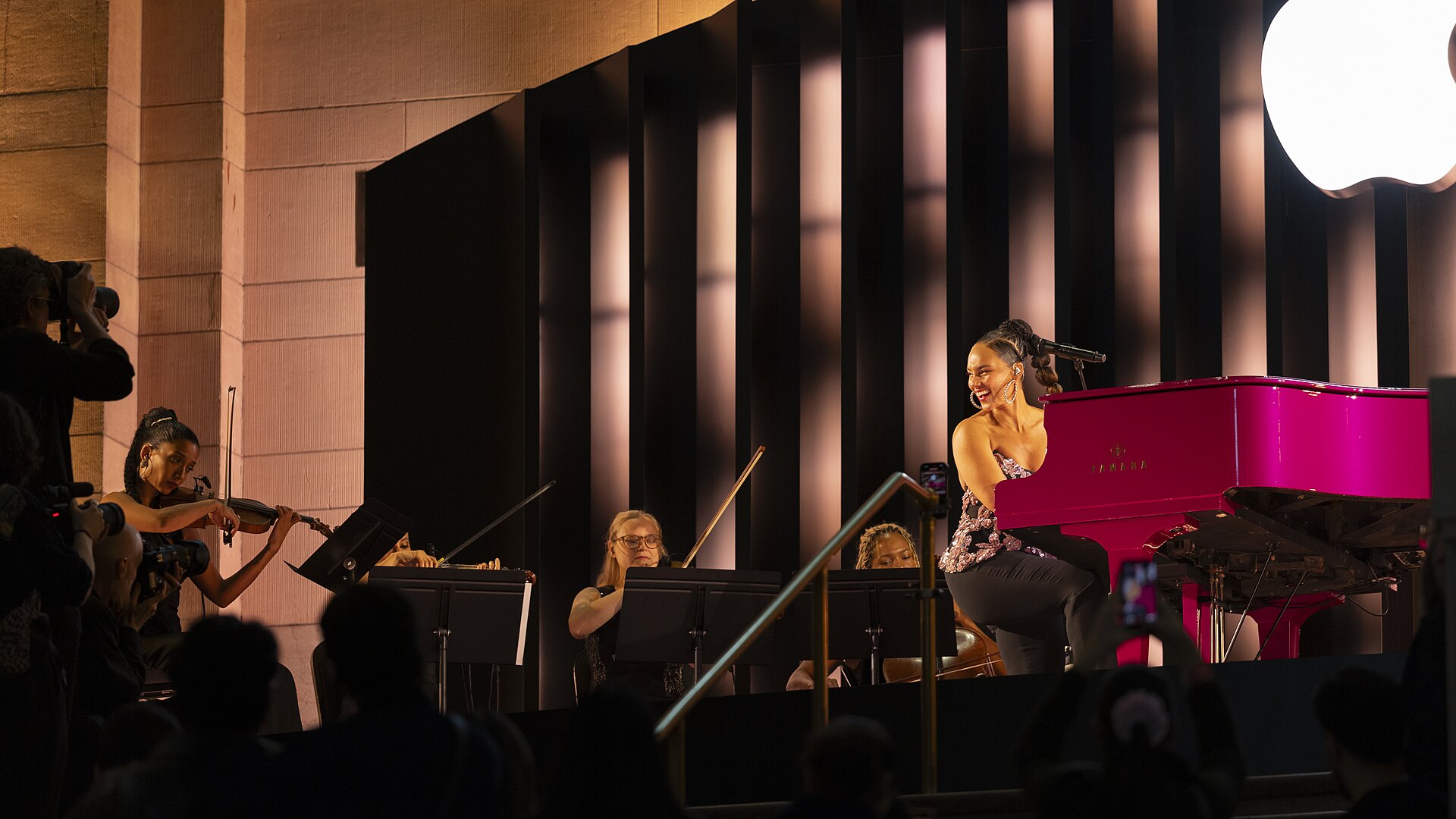 Alicia Keys performs at a bright pink Yamaha grand piano during Apple’s 50th-anniversary kickoff event at the company’s Grand Central Terminal store in New York City on 13 March 2026. There is a small accompanying string ensemble.