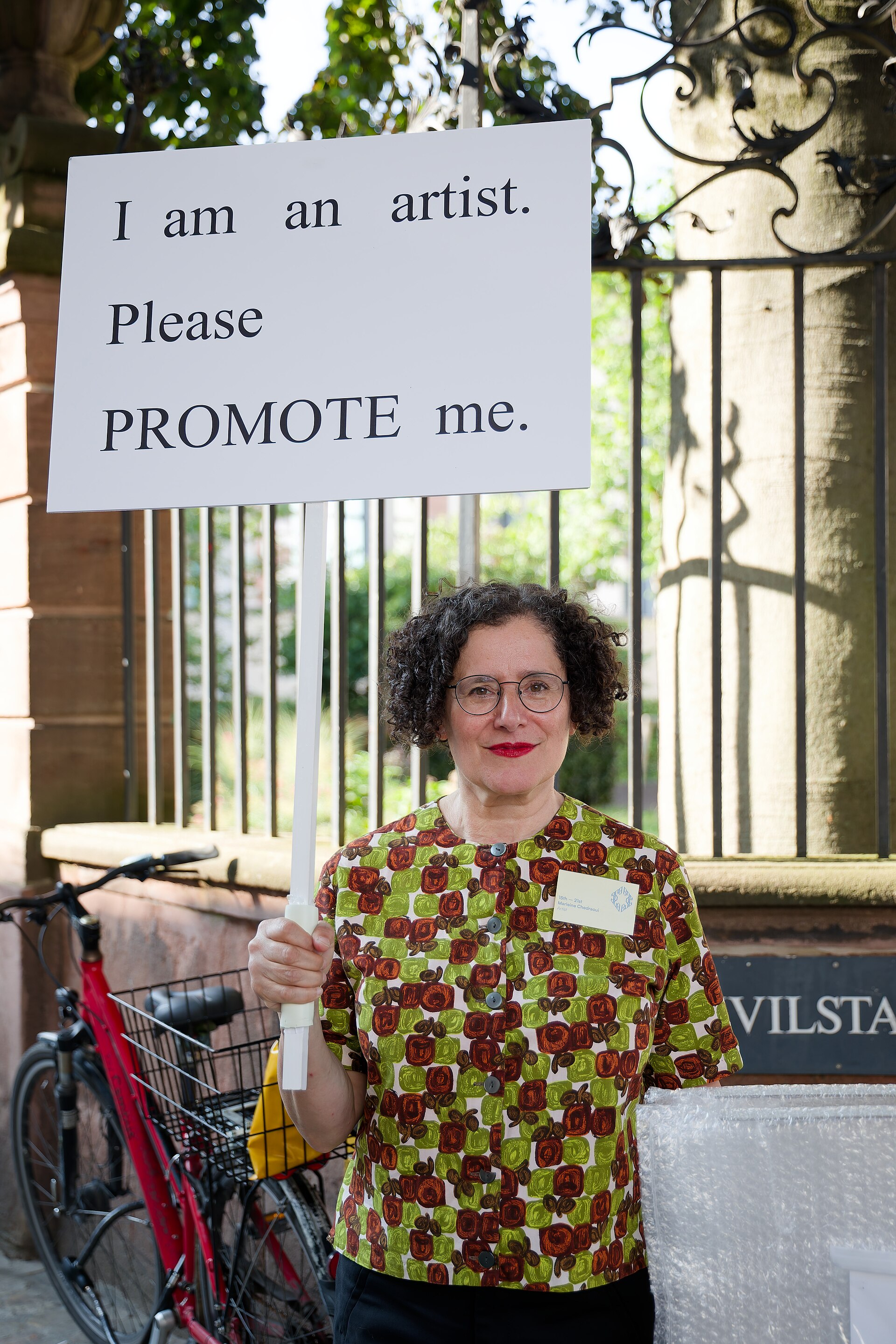 Marleine Chedraoui, artist, for the project WHILE STOCKS LAST at Basel Social Club 2025. Chedraoui holds a sign that reads, "I am an artist. Please PROMOTE me."